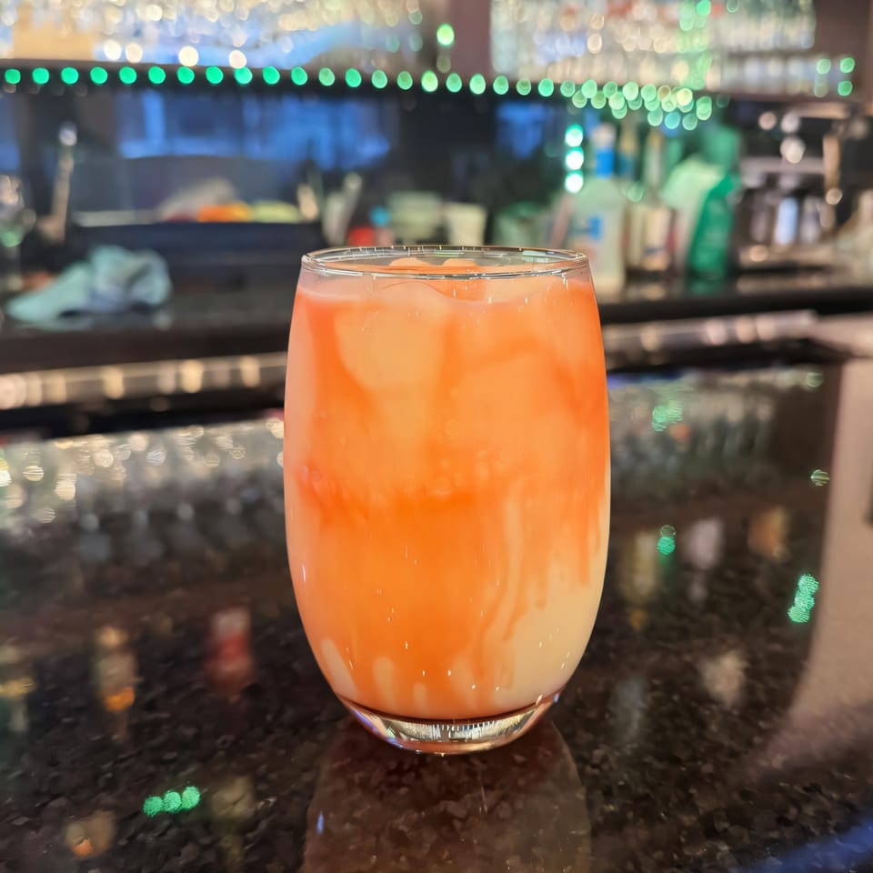 A vibrant orange and white layered drink with ice sits on a dark bar counter with blurred lights.