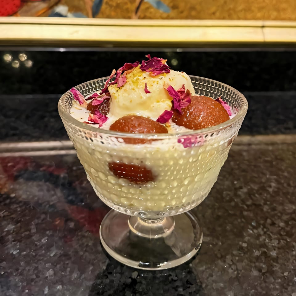 Indian dessert with gulab jamun, ice cream, and rose petals in a clear glass bowl.