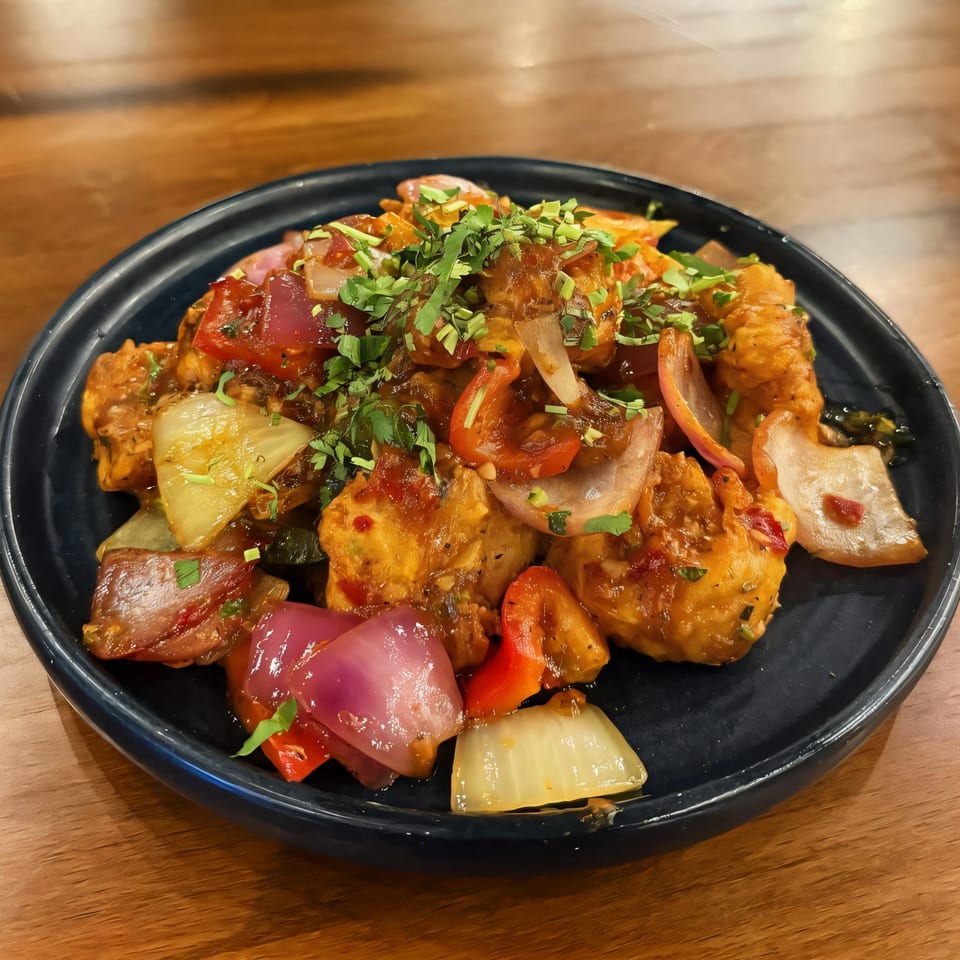 Indian chili chicken with red and white onions, bell peppers, and cilantro on a black plate.