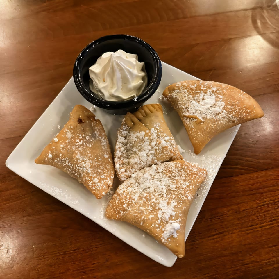 Four golden-brown Indian pastries, dusted with powdered sugar, served with a side of whipped cream.
