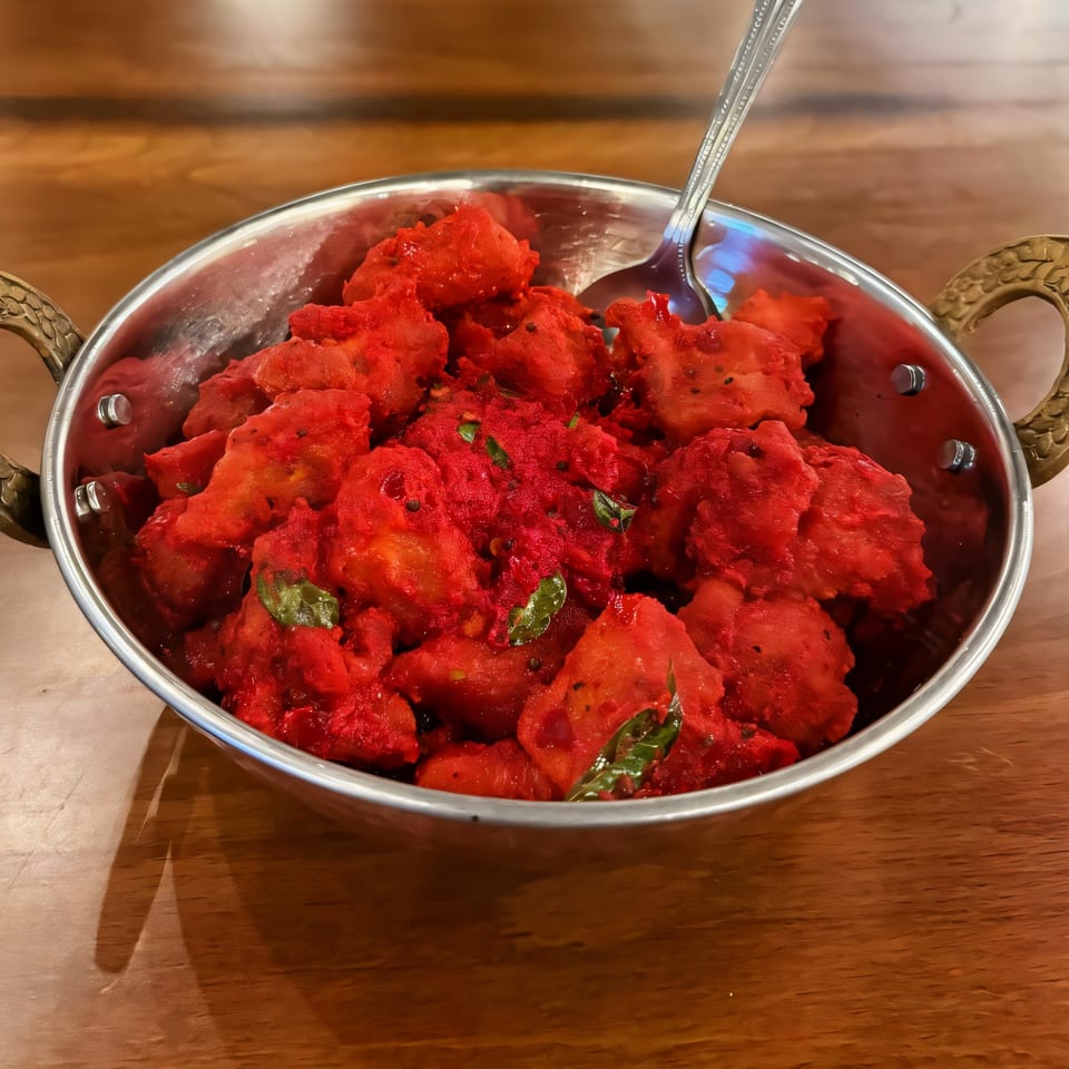 Bright red Indian chicken 65, garnished with curry leaves, in a metal serving bowl with a spoon.
