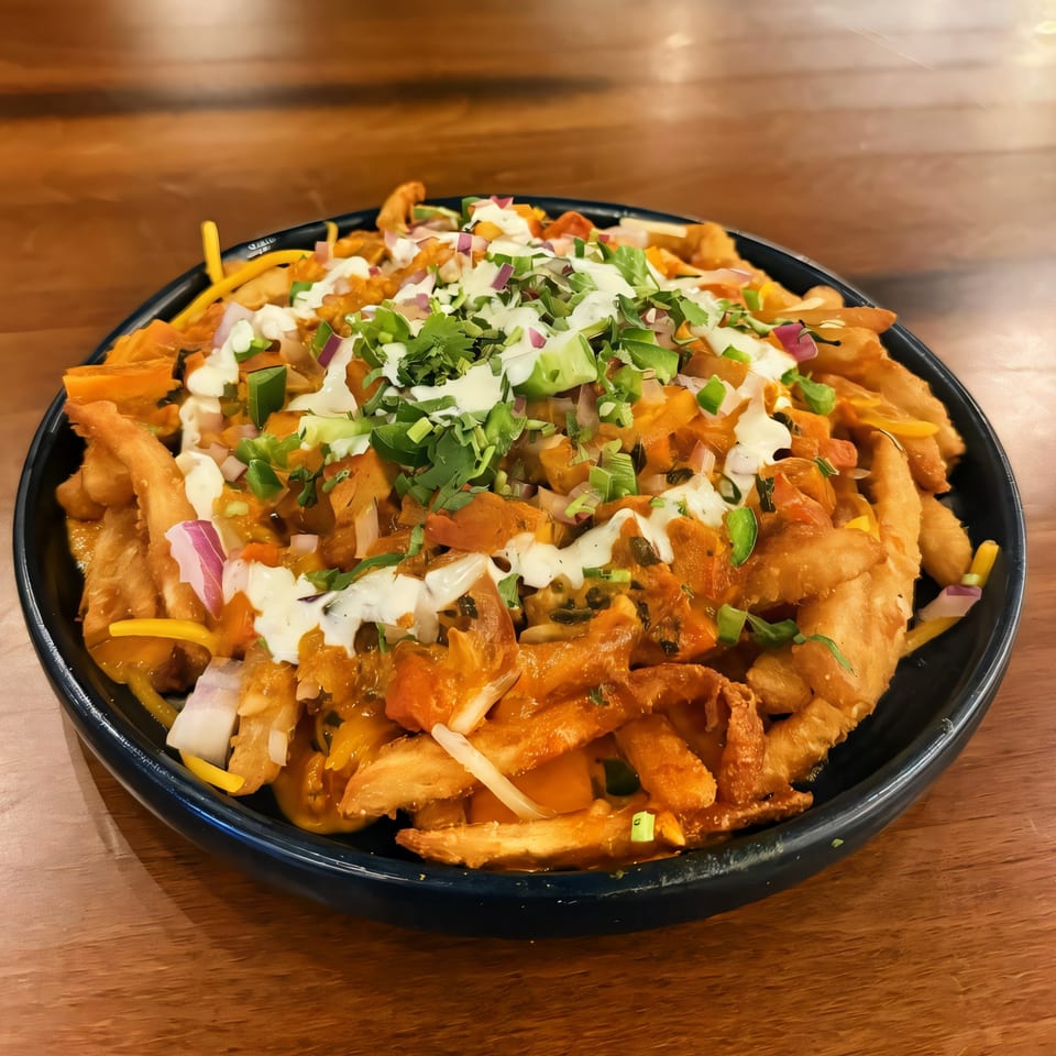 A black plate of Indian-style loaded fries with orange sauce, white drizzle, and fresh herbs.